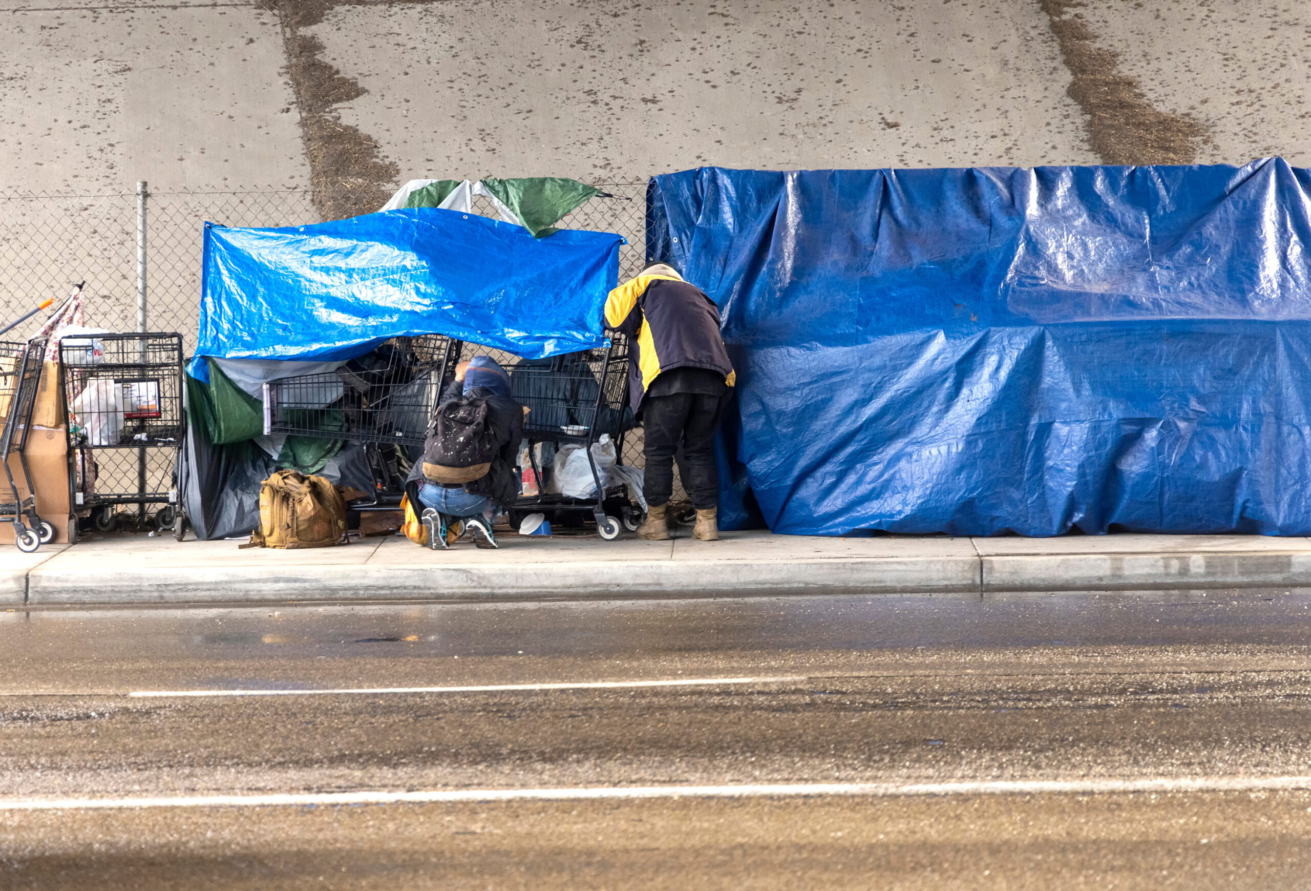 Homeless people at a camp with blue tarp and a car passing in th